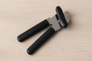 A manual can opener with black handles rests on a light wooden surface.
