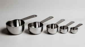 A set of six stainless steel measuring cups, essential baking tools, arranged in a row from largest to smallest on a white surface—a perfect addition to any kitchen utensils set.